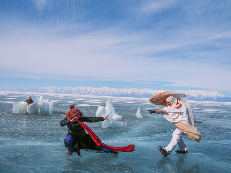 Two performers are facing each other on the frozen sea with ice caps behind them. One performer is low to the ground with their arm stretched twoards the other. The other performer is pointing their gloved finger towards the first performer.