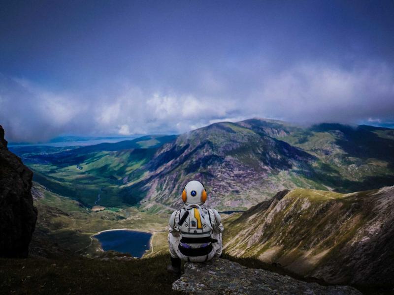 A person in a spacesuit sitting on a mountain looking out over green mountains with a dark blue lake in the distance and a cloudy sky above.
