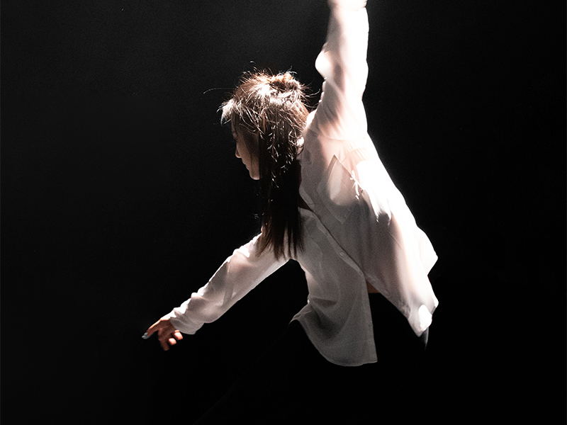 A person wearing a white shirt and black trousers is dancing on dark stage. There is bright light focused on the dancer. Their gaze, and right arm is outstretched towards their right foot whilst their left arm is reaching to the opposite direction.
